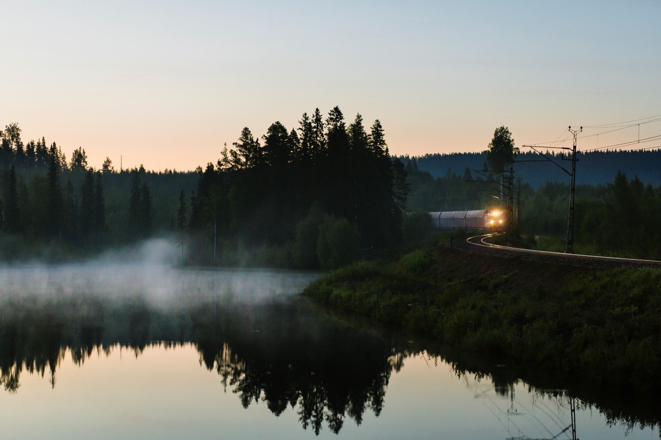 Fotografi på solnedgång vid sjö, ett tåg passerar och dimma ligger över sjön.